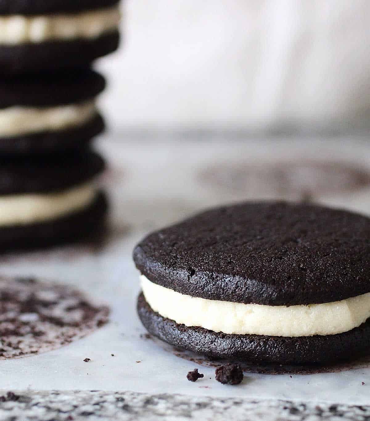 Single dark chocolate homemade Oreo cookie on parchment paper; stack of cookies in the background.