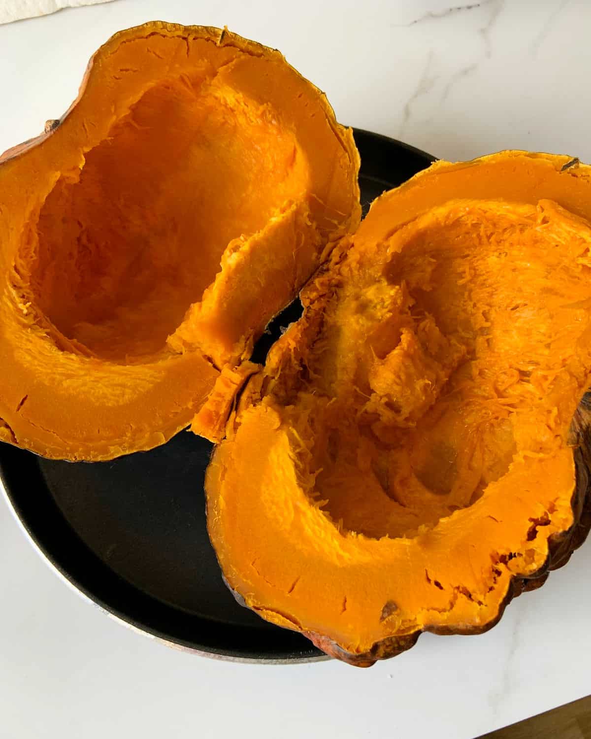 Opened baked pumpkin without seeds on a dark round pan on a white surface.