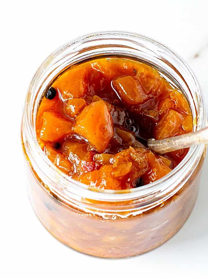 Glass jar with peach chutney and a silver spoon. White marble surface.
