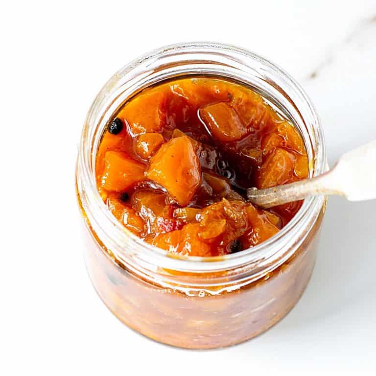 Glass jar with peach chutney and a silver spoon. White marble surface.