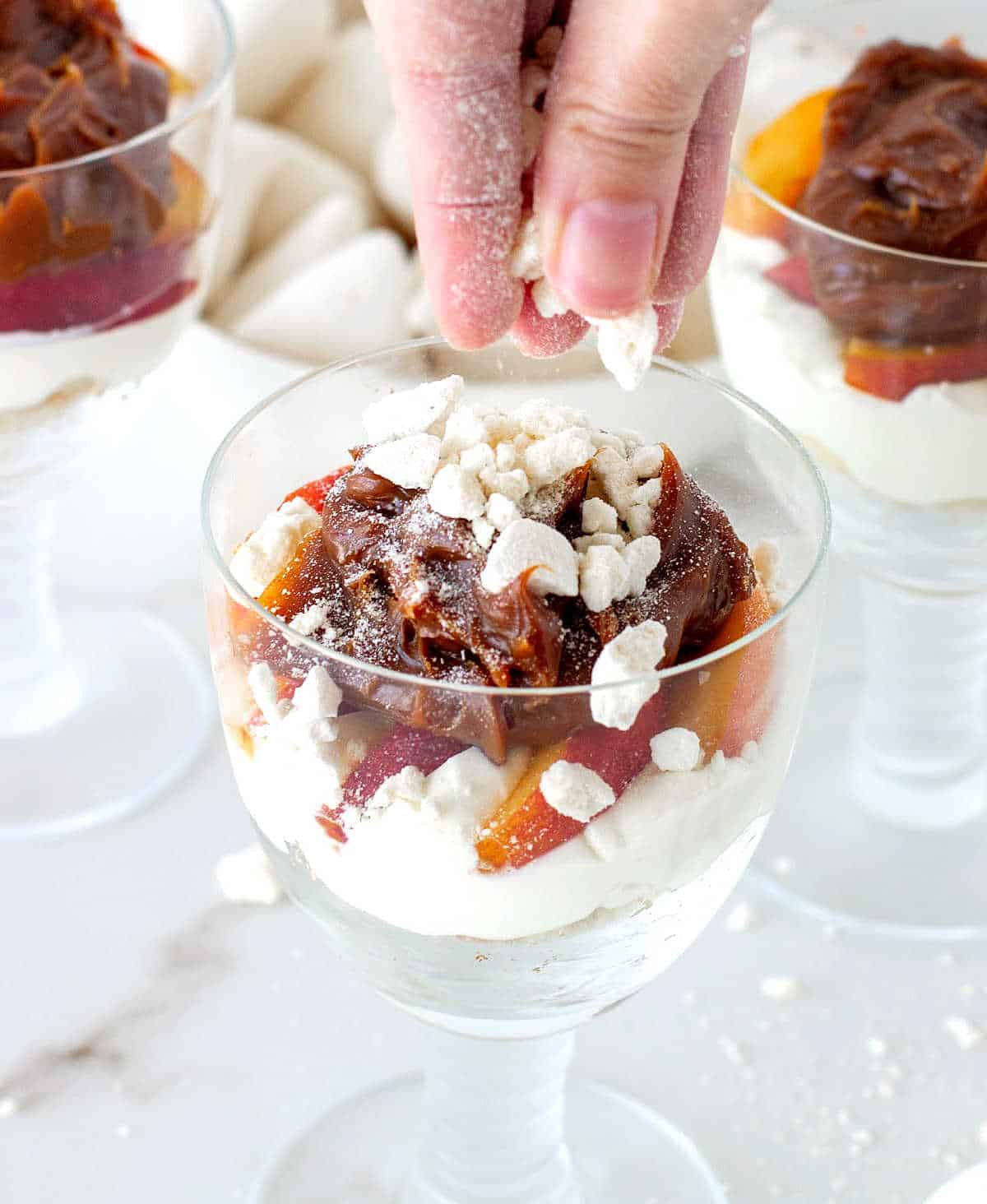 Crushed meringue added to glasses containing peach cream trifle on a white marble table.