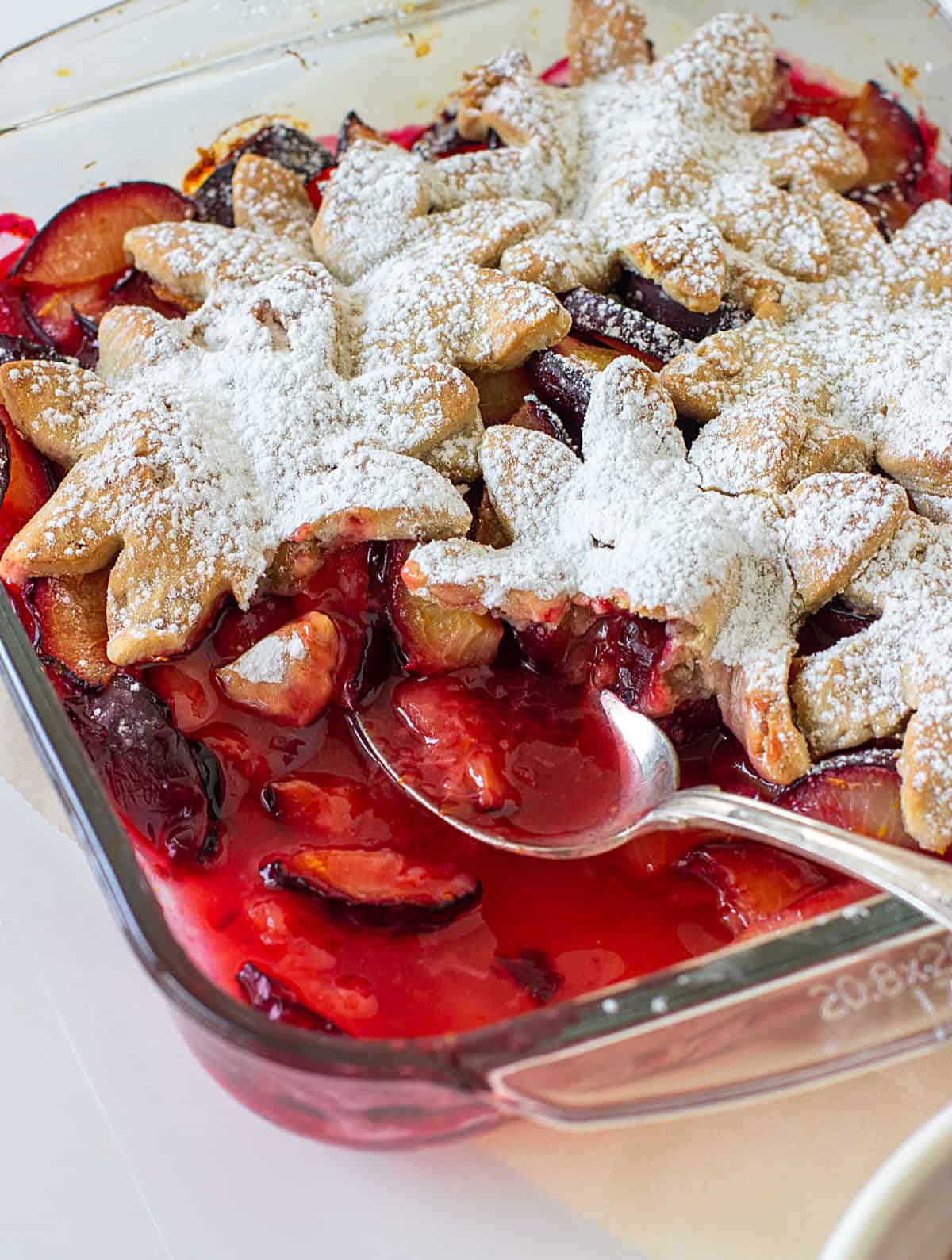 Glass dish with plum cobbler made with a pie crust topping, a silver spoon inside 