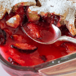 Glass dish with plum cobbler, silver spoon inside, pie dough cut-outs as topping. Image with text