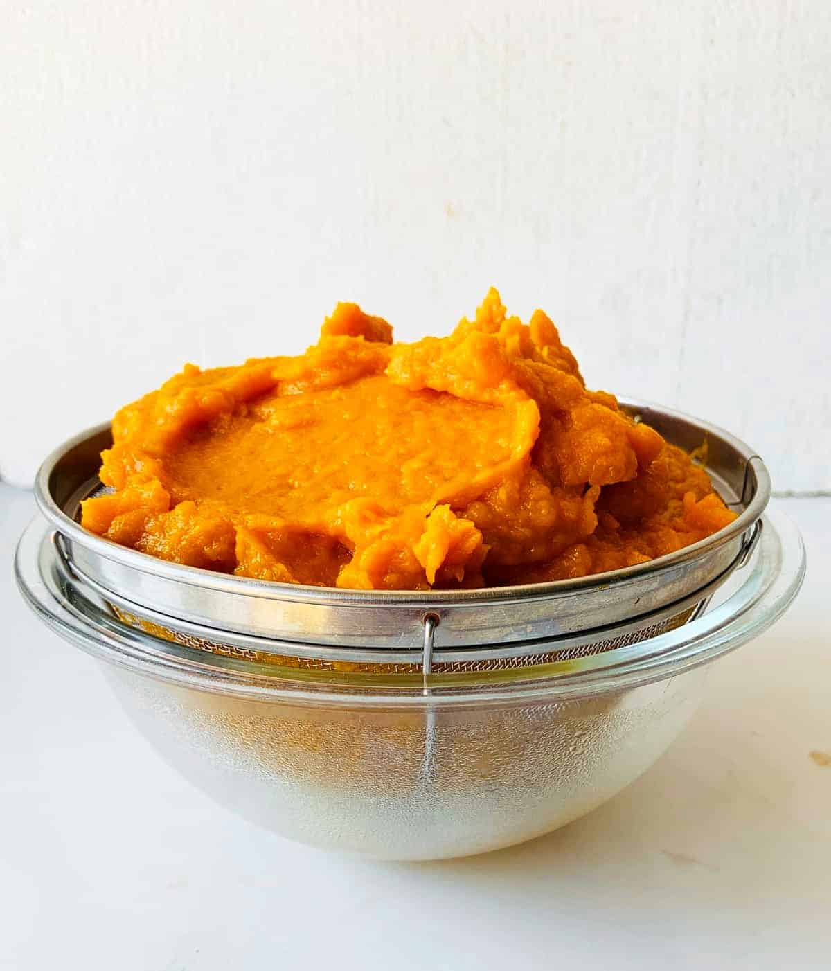 Colander with pumpkin puree over a glass bowl. White surface and background.