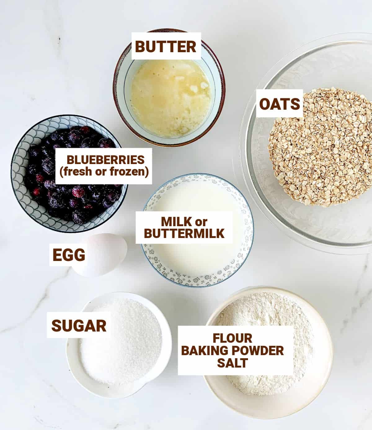 Different bowls with blueberry oatmeal muffins ingredients on a white surface.