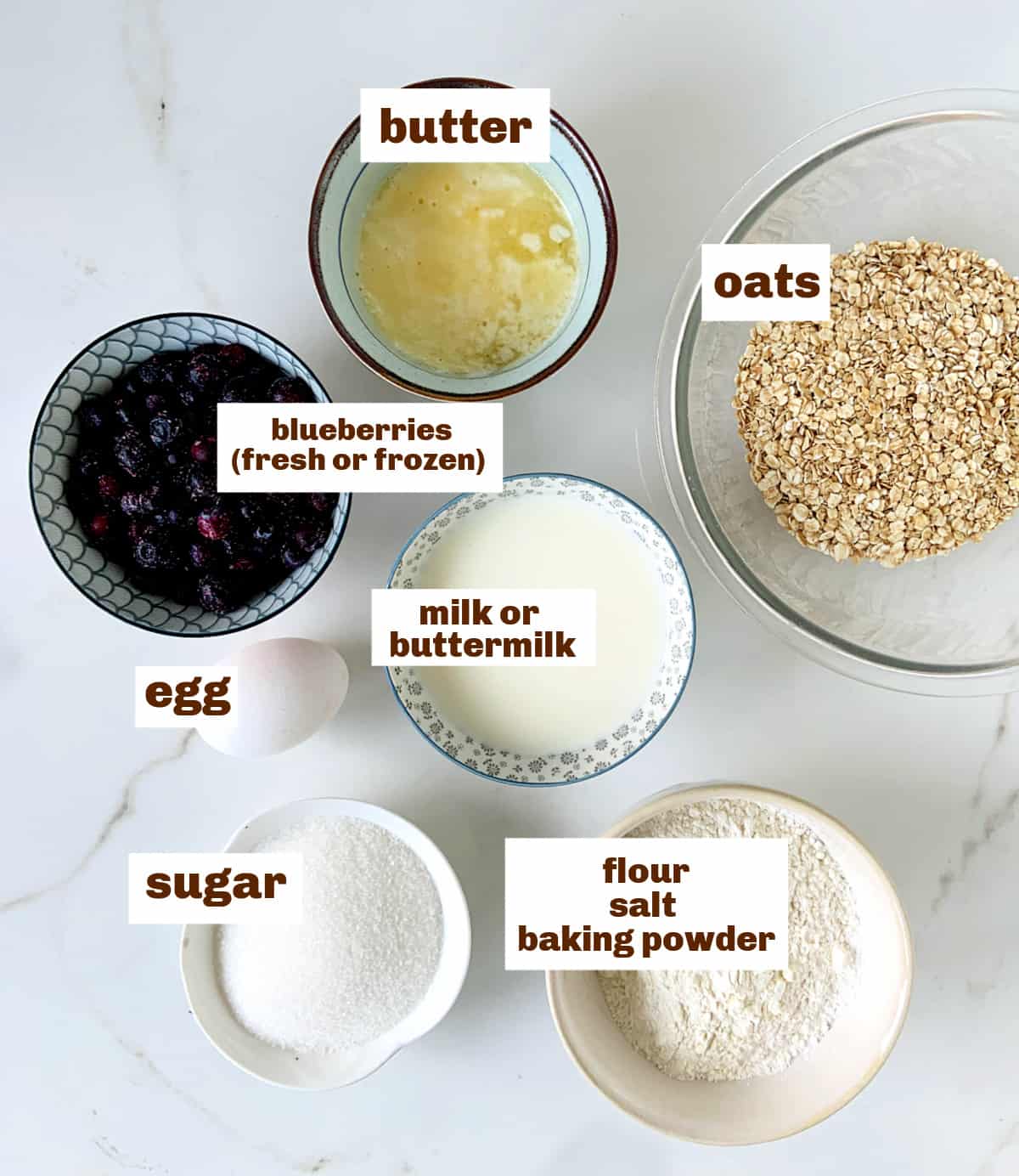 Different bowls with blueberry oatmeal muffins ingredients on a white surface