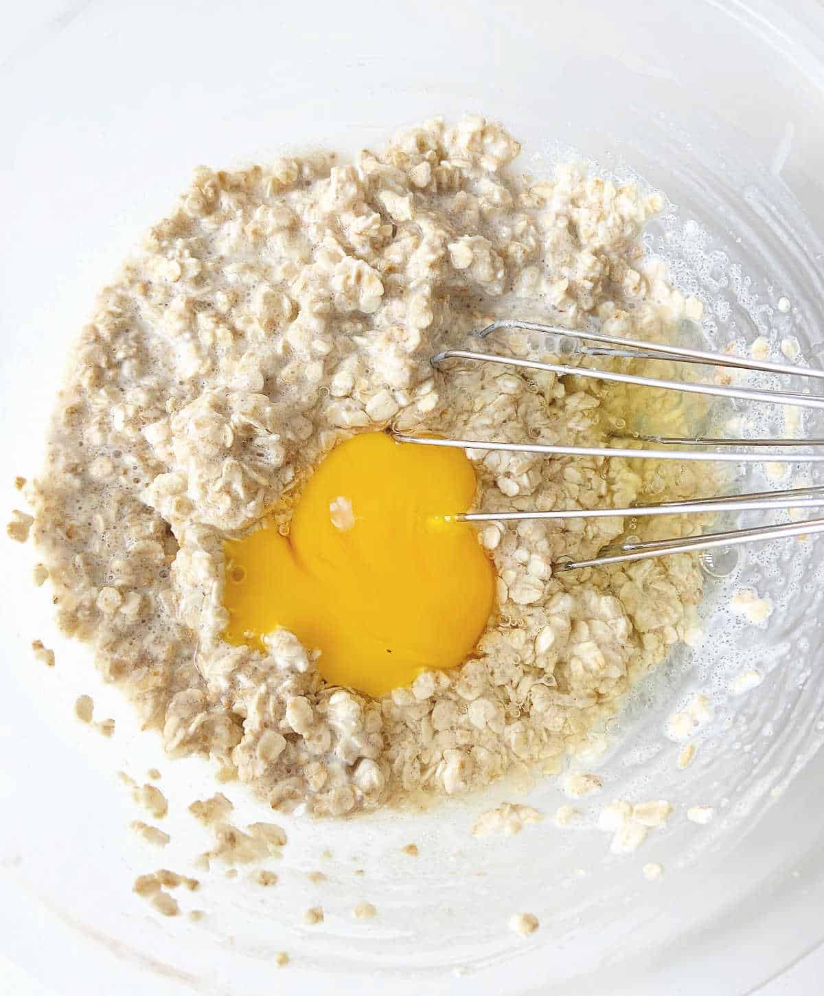Eggs added to oatmeal muffin mixture in glass bowl with a whisk inside.