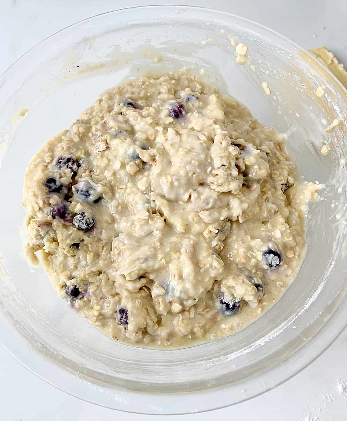 Mixed muffin batter with blueberries in a glass bowl on a white surface.