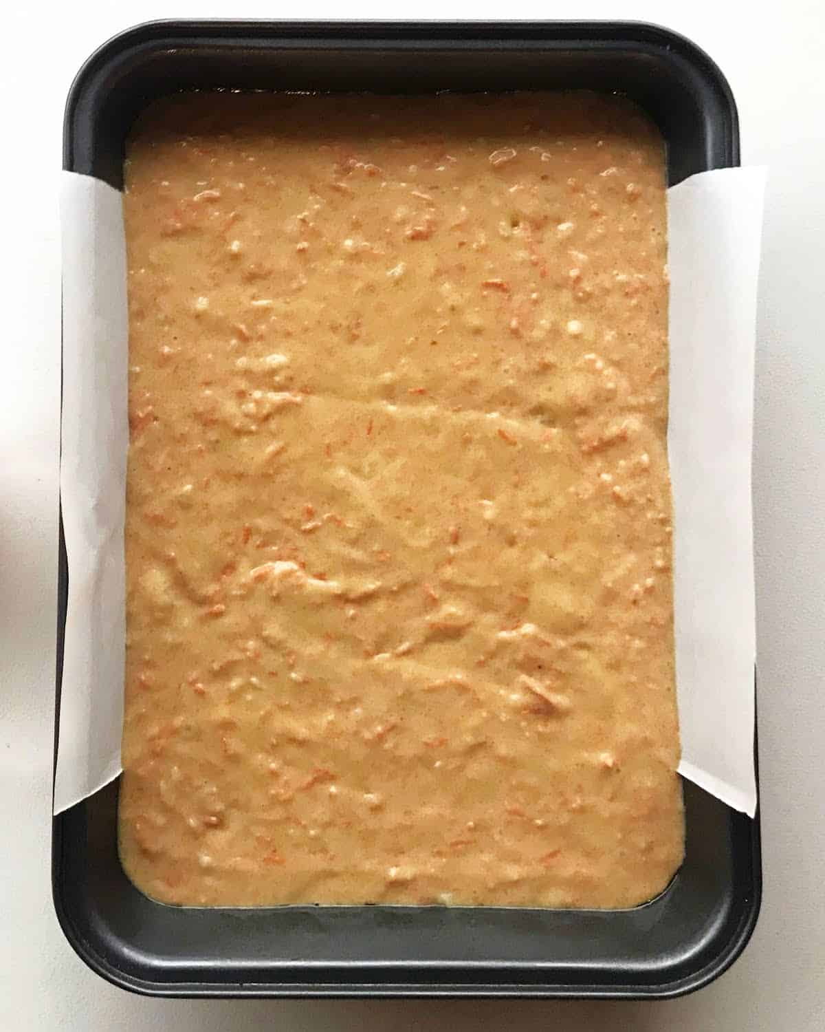 Carrot cake batter in a dark metal rectangular cake pan with parchment paper. White surface.