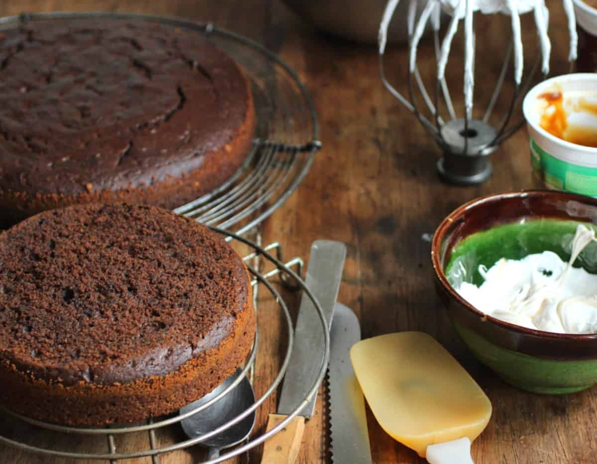 Wooden tables with baked chocolate cakes on wire racks, bowl and beater with meringue, utensils.