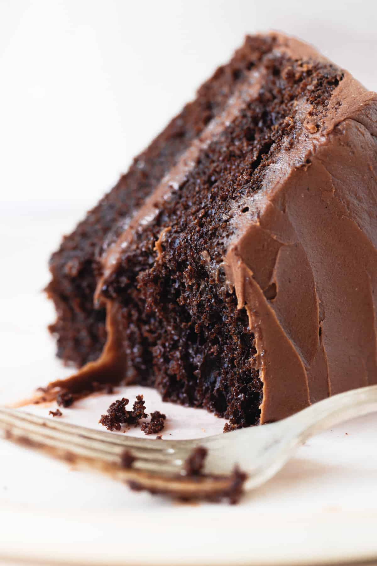 A slice of eaten frosted chocolate layer cake on white plate and background, a silver fork
