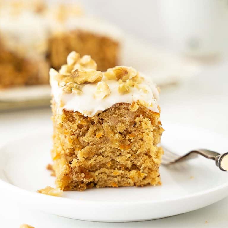 Frosted carrot pineapple cake square sprinkled with walnuts on a white plate with a fork. More cake in the white background.