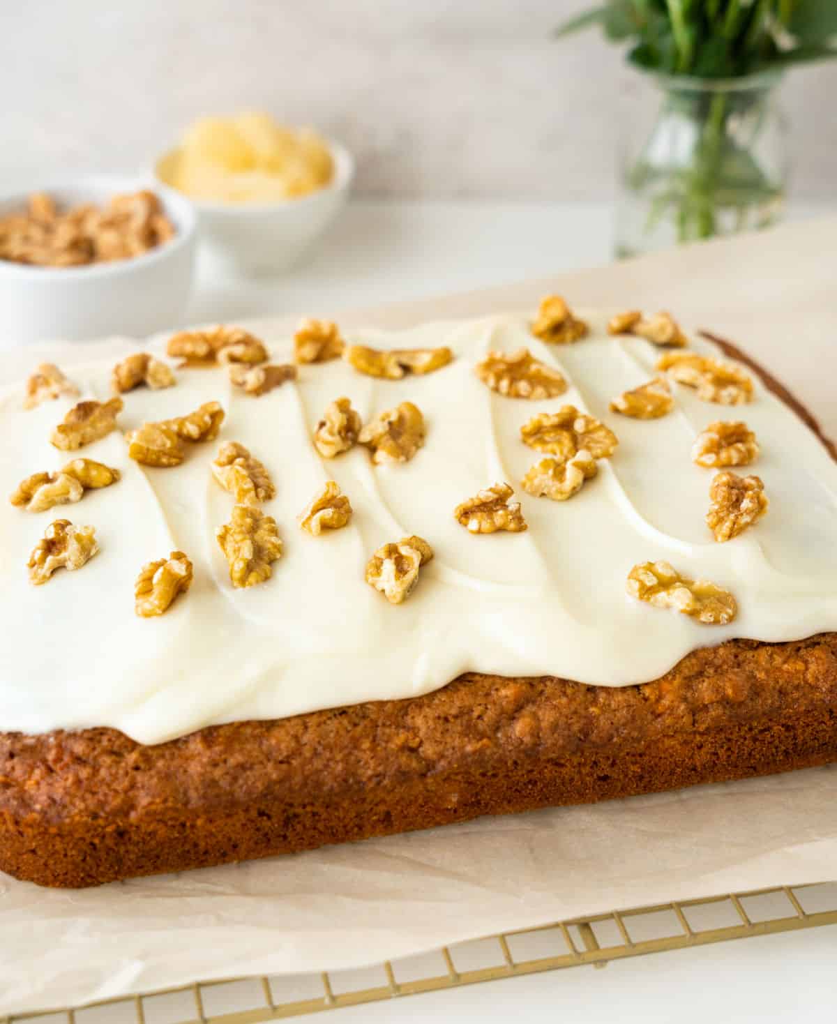 Frosted whole carrot sheet cake on a beige paper with bowl and ingredients on a grey background.