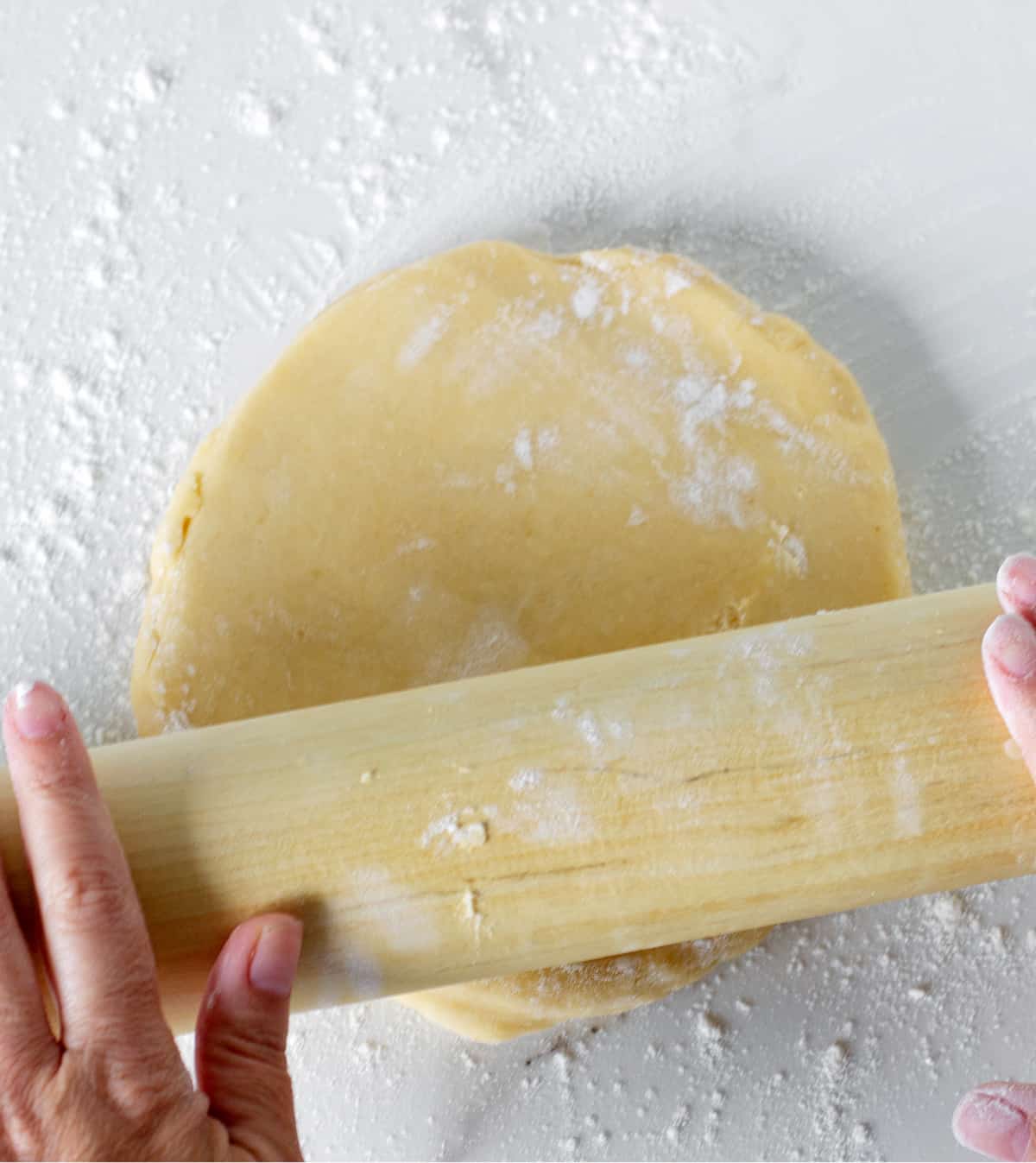 Hands rolling disc of dough with pin on white surface.