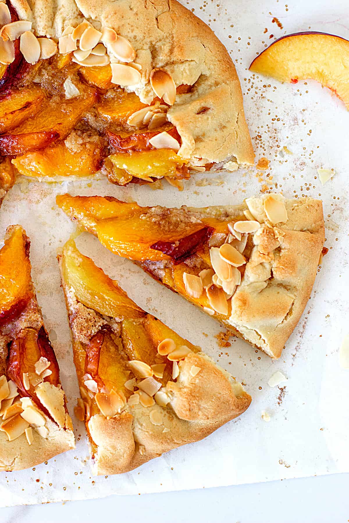 Two cut slices of a peach galette and rest of tart on a white surface, fresh peach slice