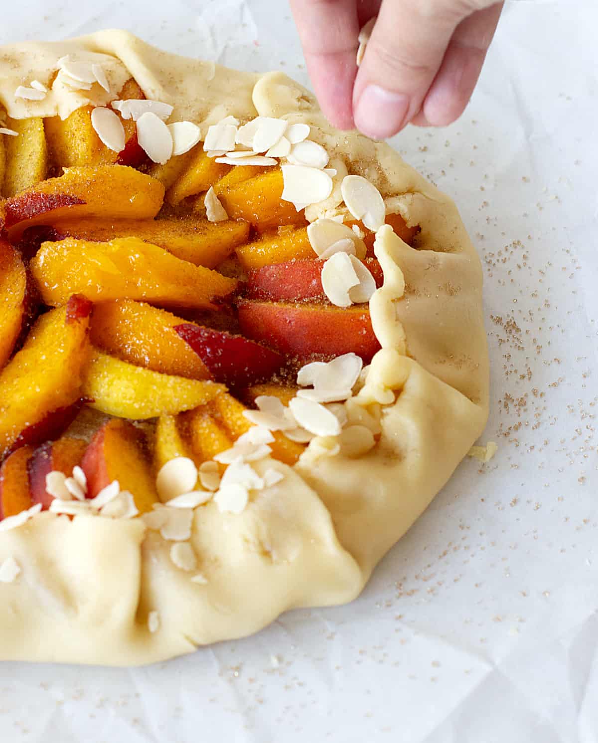 On a white surface an unbaked peach free-form tart, hand adding sliced amonds.