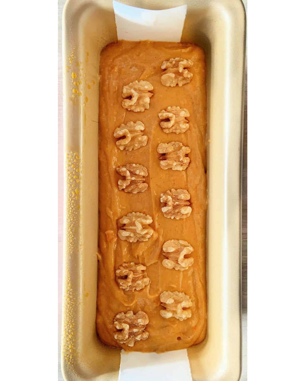 Pumpkin bread batter with walnuts on a loaf pan on a white surface.