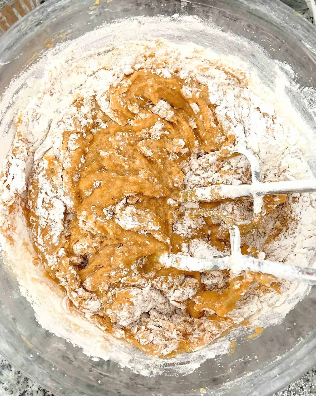 Flour added to pumpkin bread batter in a glass bowl with metal beaters inside.