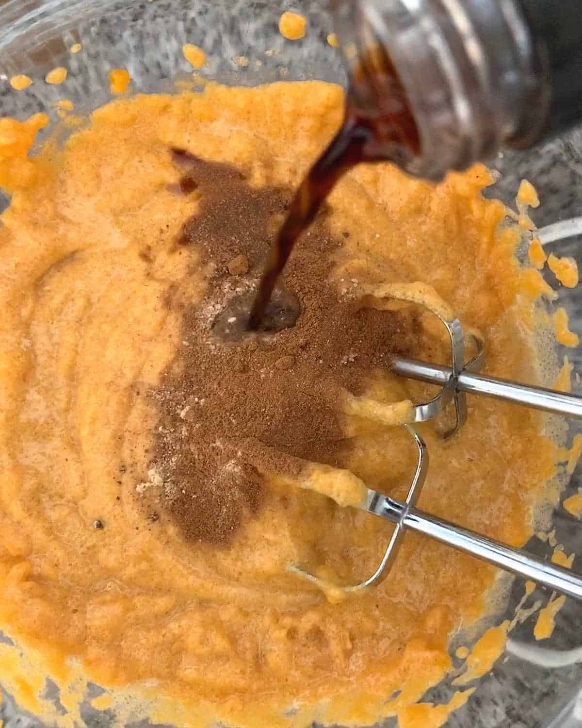 Adding vanilla and spices to pumpkin bread batter in a glass bowl with metal beaters.