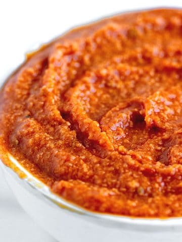 Close up of reddish orange romesco sauce in a white bowl on a white background.