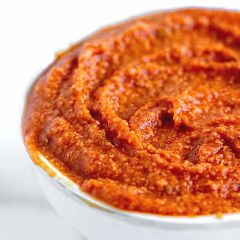Close up of reddish orange romesco sauce in a white bowl on a white background.