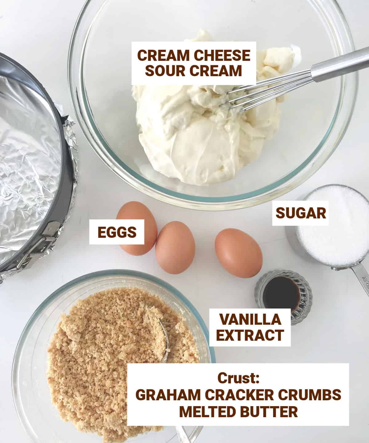 White surface with bowls with ingredients for cheesecake including eggs, sugar, crust mixture, sour cream, vanilla. Text overlay.