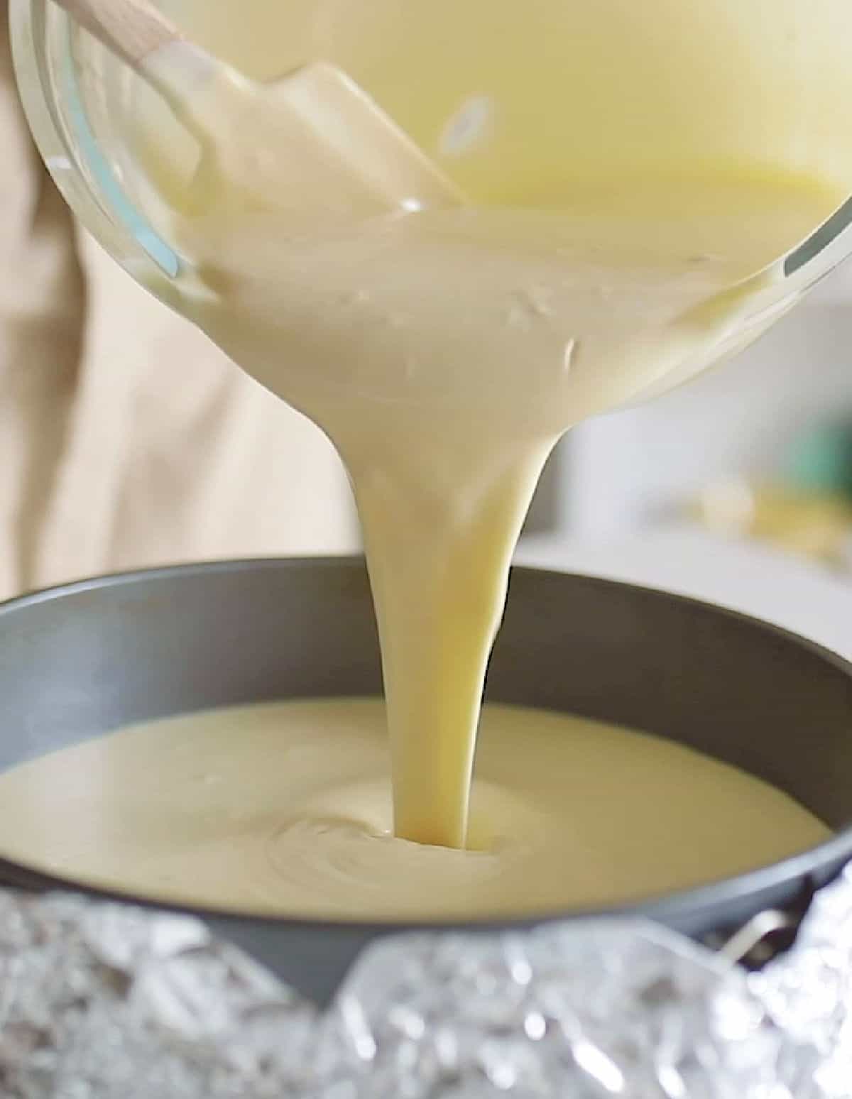 Pouring cheesecake mixture into round cake pan lined with aluminum foil.