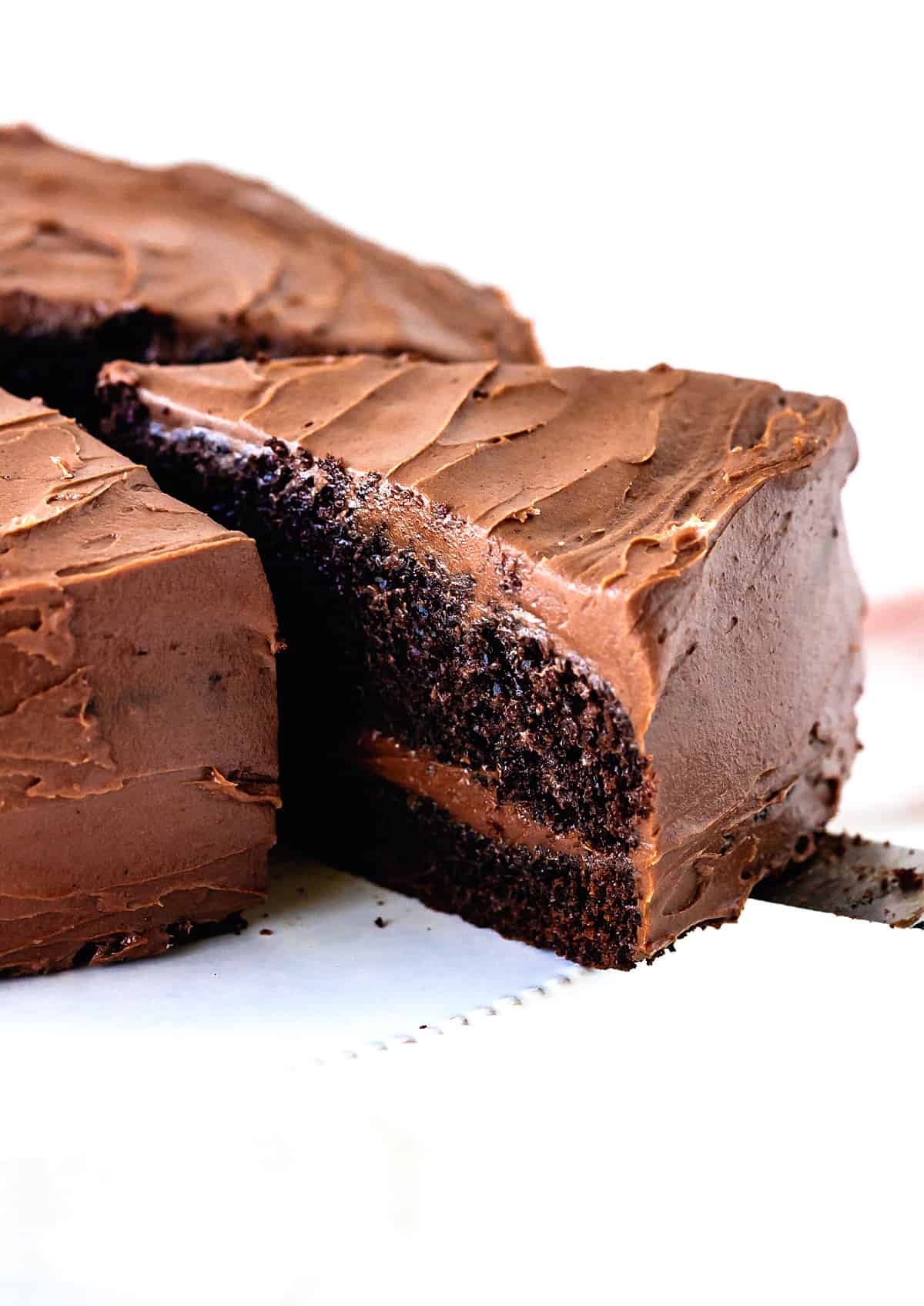 A slice of frosted two layer chocolate cake being pulled away from whole cake on white cake stand.