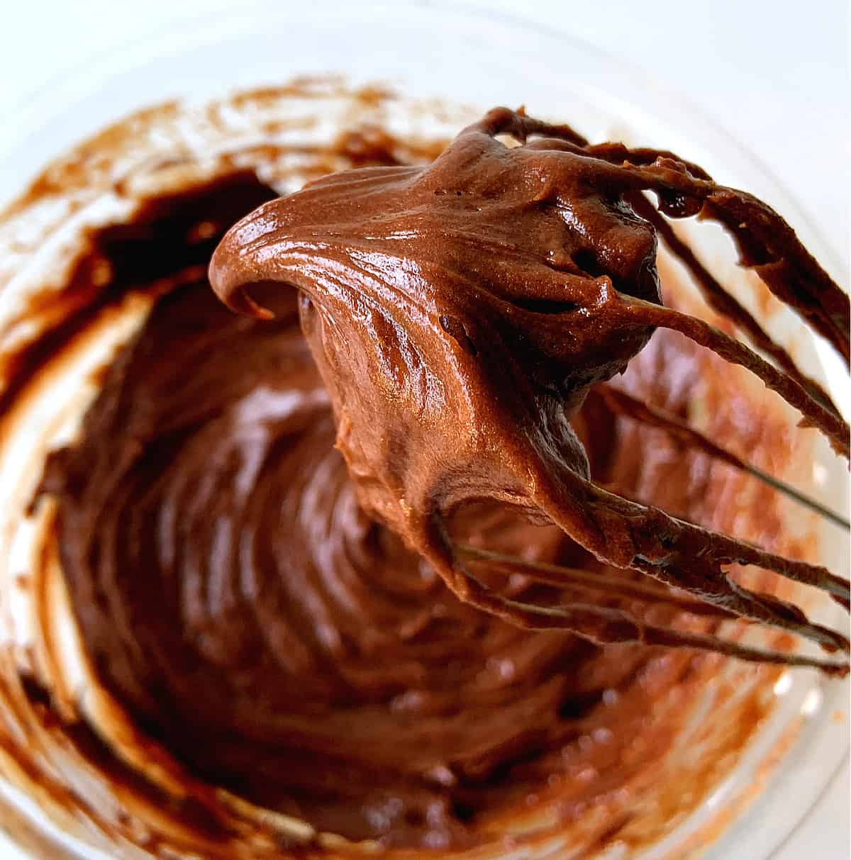 Wire whisk with dollop of chocolate frosting, glass bowl with rest of mixture below