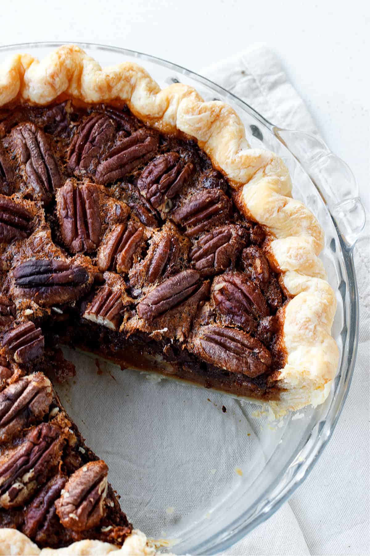 Partial view of pecan pie in glass dish on white table, one slice missing