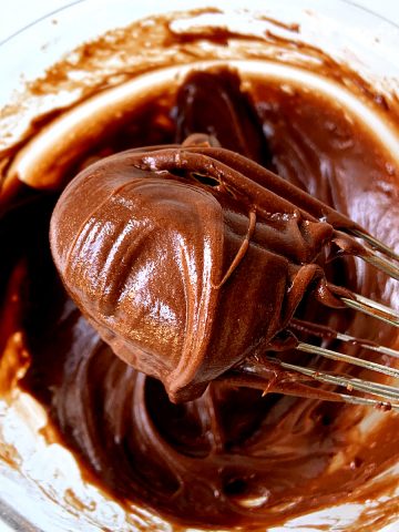Shiny dollop of chocolate frosting on a wire whisk, bowl with more mixture below