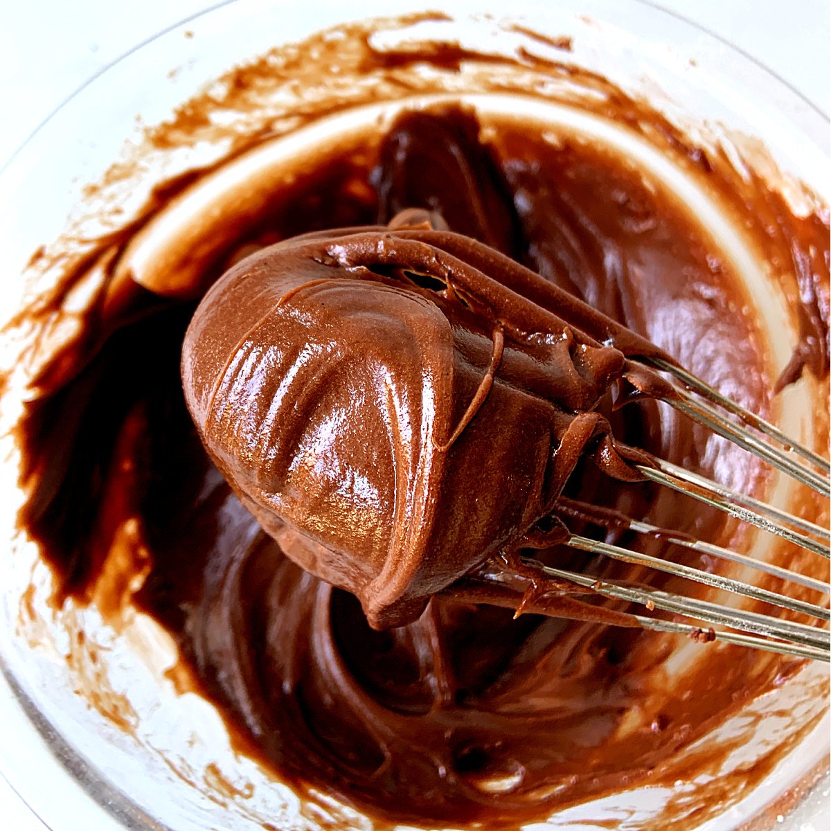 Shiny dollop of chocolate frosting on a wire whisk, bowl with more mixture below.