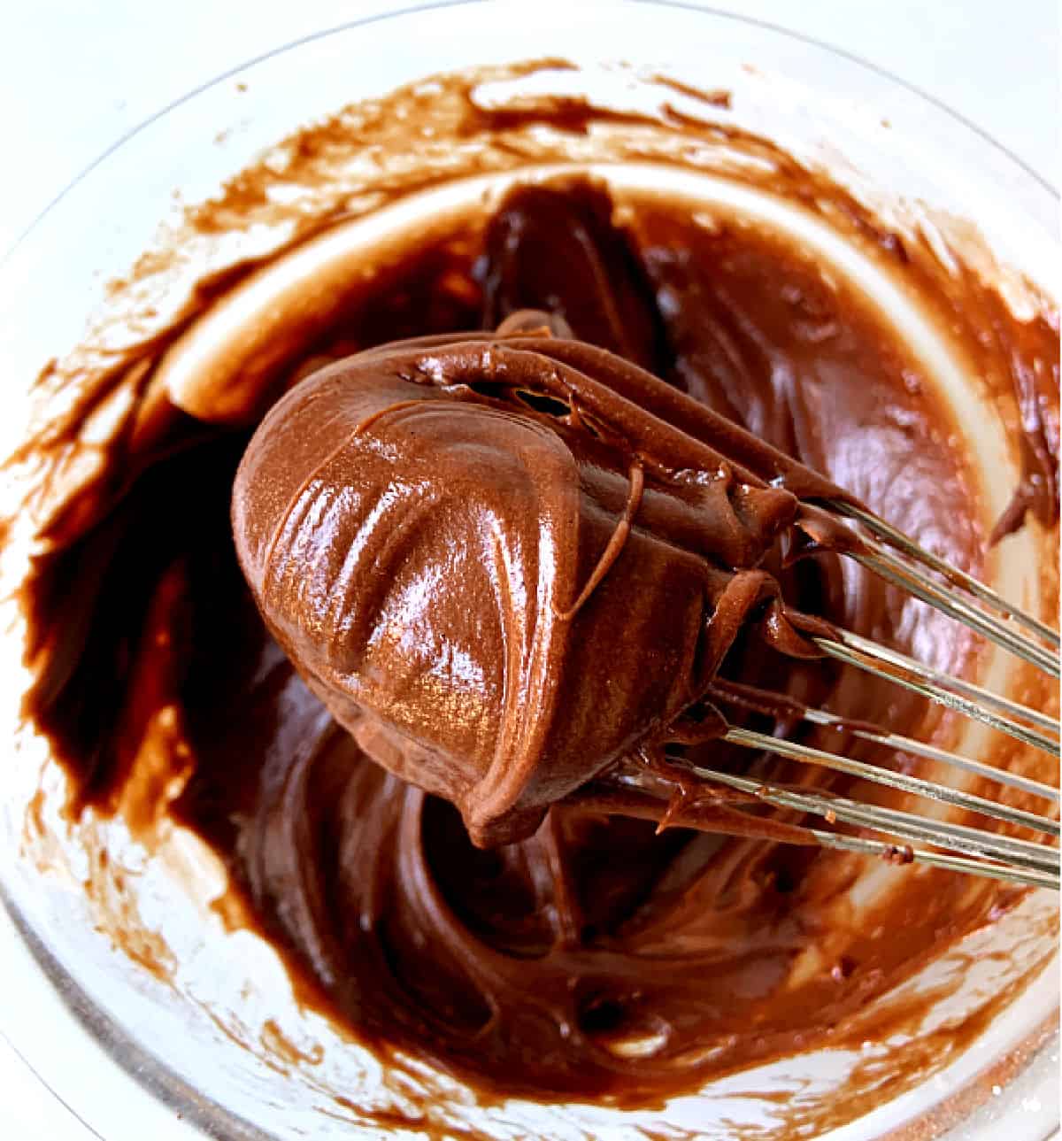 Close up of smooth shiny chocolate frosting on a wire whisk, rest of frosting in glass bowl below