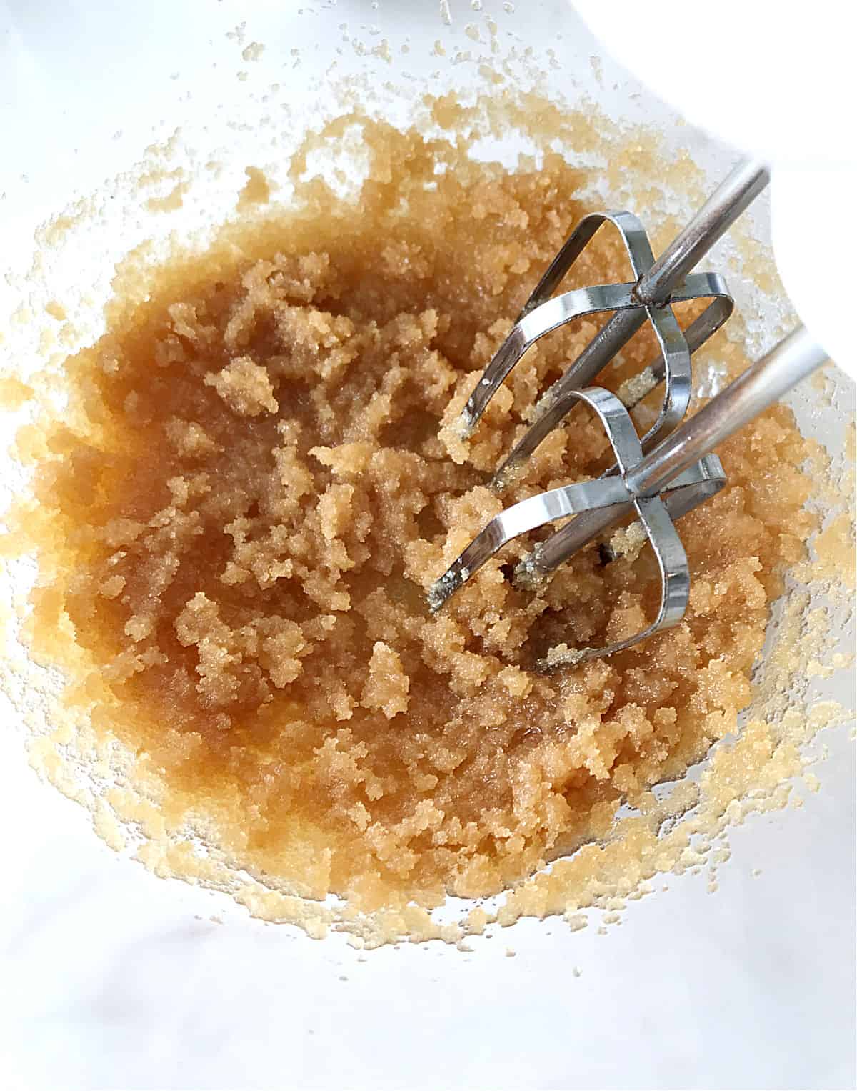 Metal beaters mixing oil and brown sugar in glass bowl, white surface