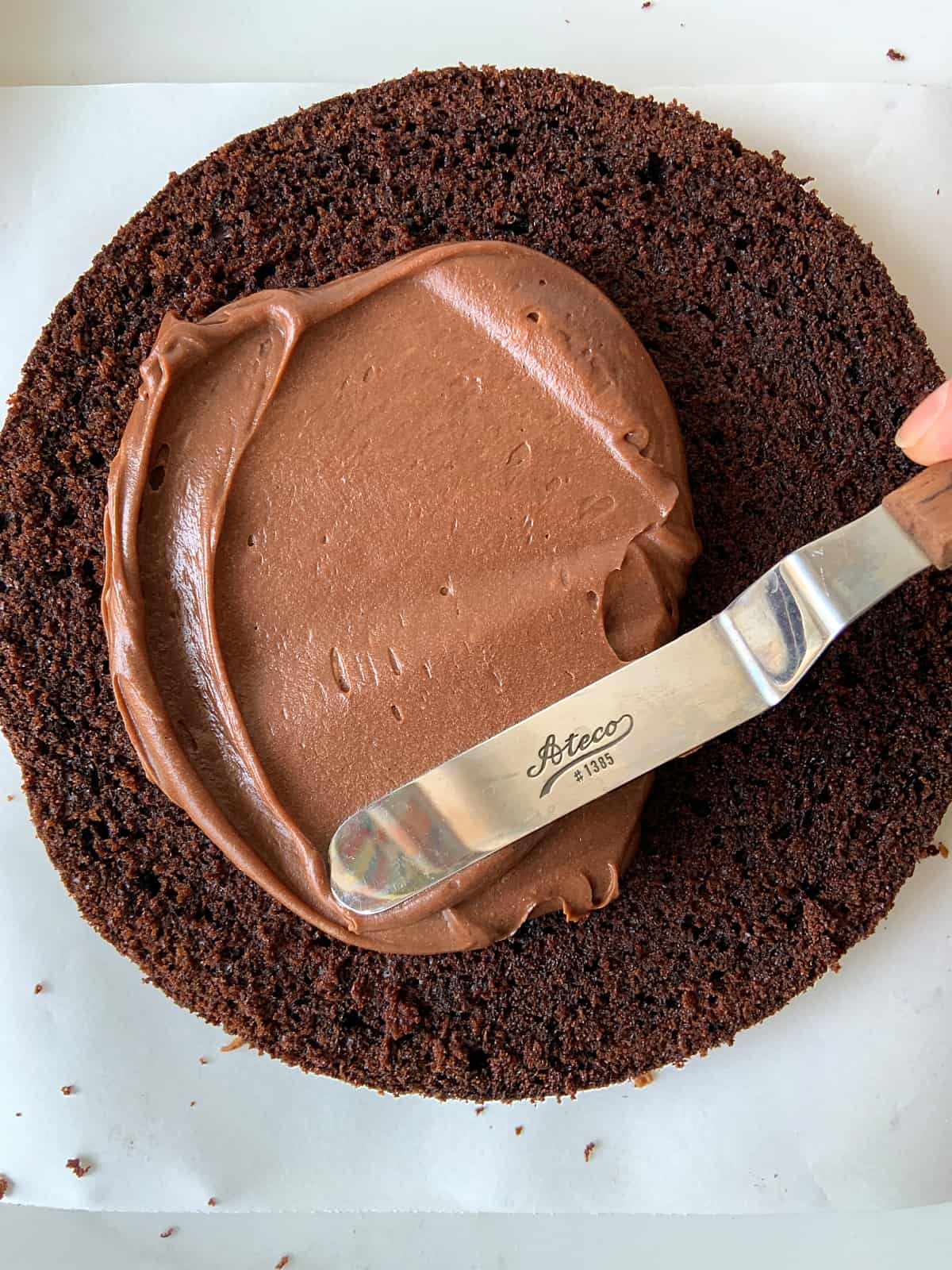 Spreading chocolate frosting with a metal spatula on a round layer of chocolate cake, white surface.