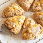Several triangle glazed scones with orange zest on white paper, metal cake server underneath