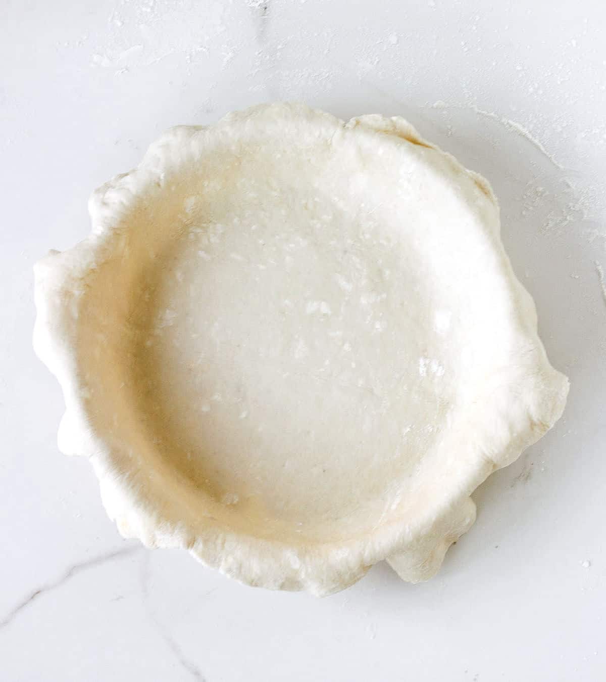 White surface with pie dough overhanging on top of a pie dish.