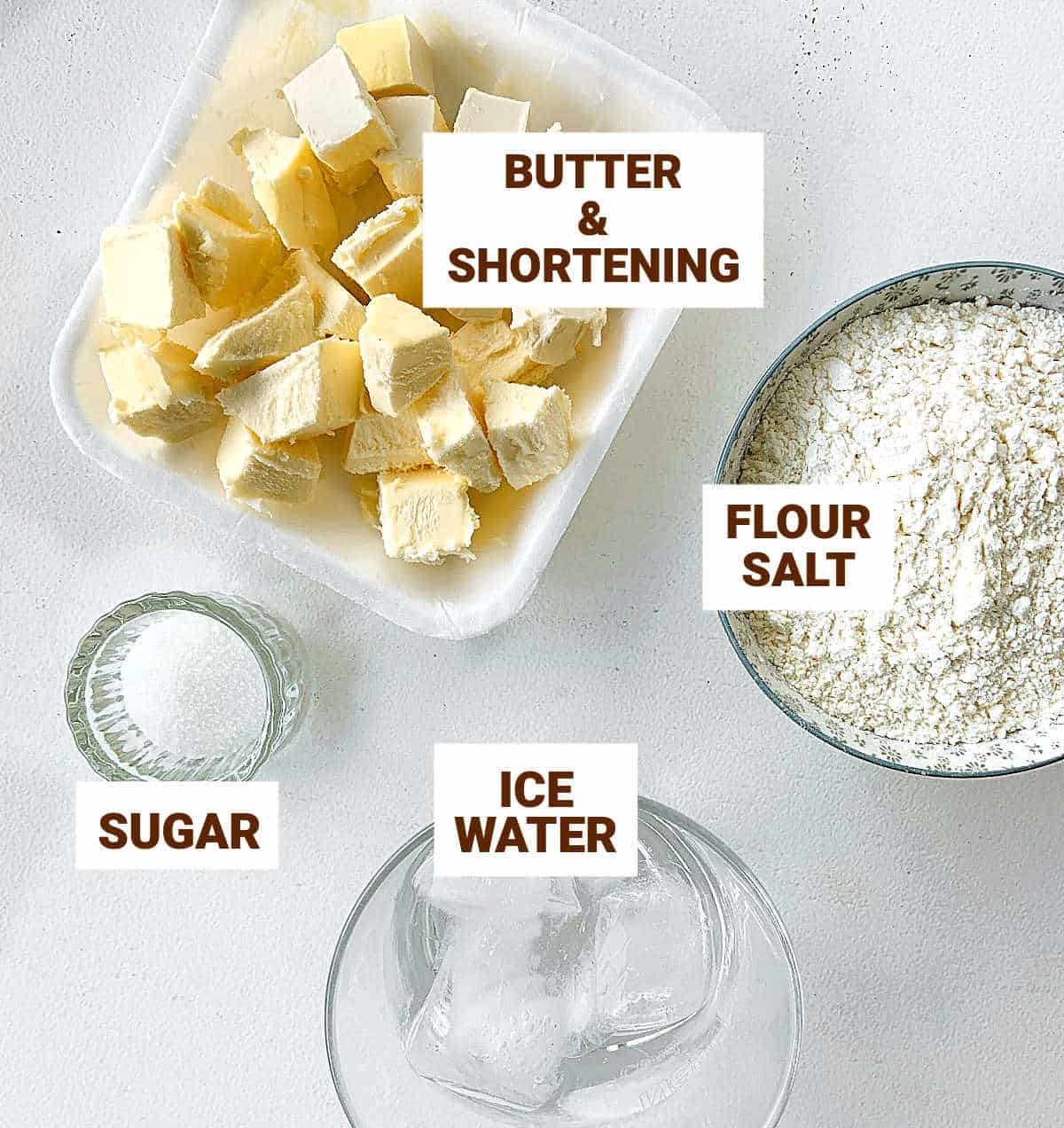 Ingredients for pie crust in bowls on a white table including butter, flour, water, sugar.
