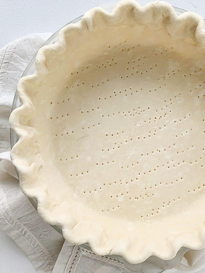 Partial view of unbaked pie crust with crimped edges on a white linen and table
