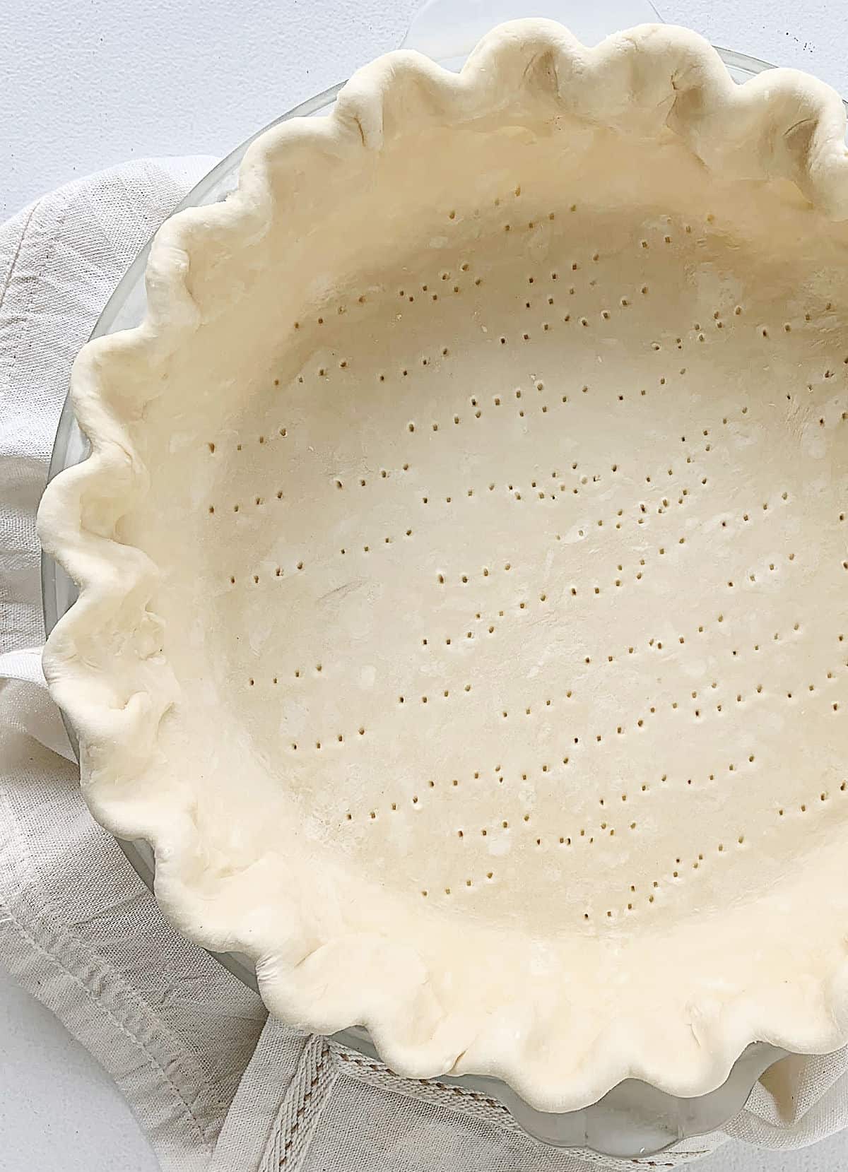 Partial view of unbaked pie crust with crimped edges on a white linen and table.