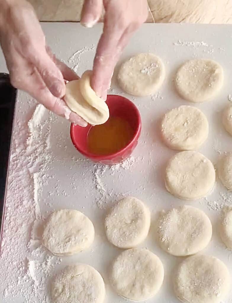 Top view of bread dough rounds on a white table. Hands forming parker house rolls. Red bowl with butter.