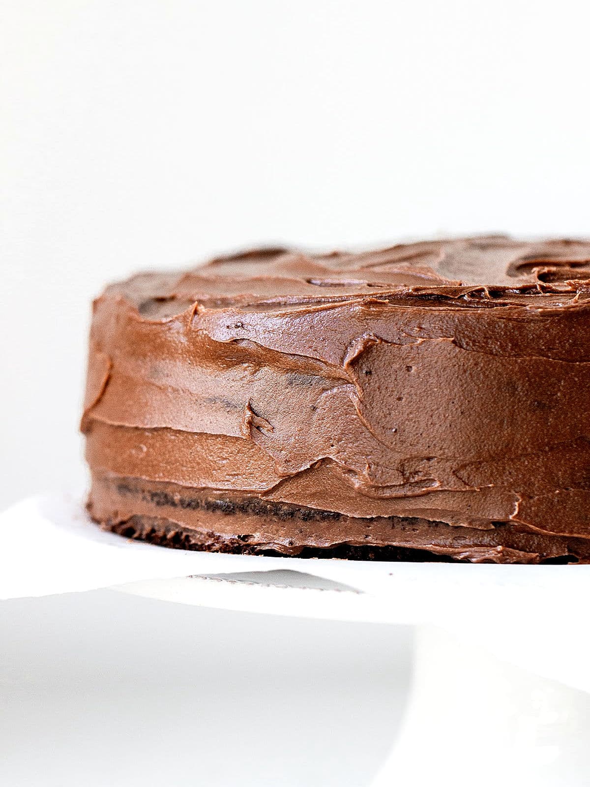 Partial view of frosted chocolate cake on a white cake stand with a white background.
