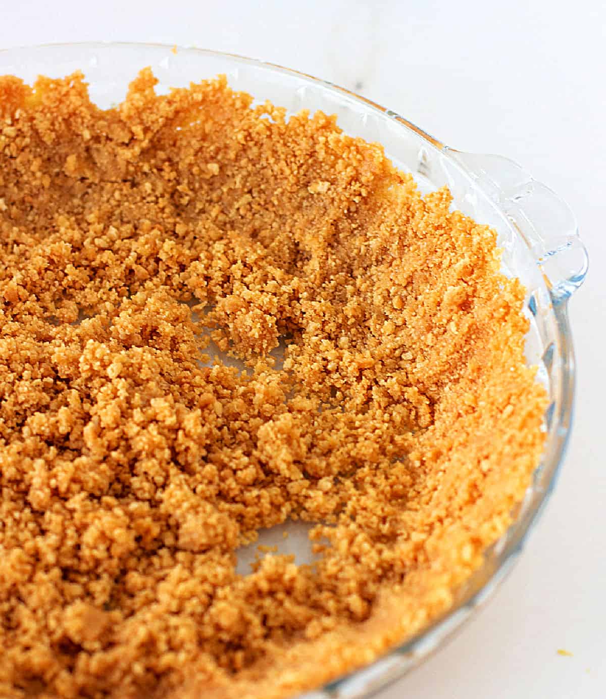 Graham cracker cookie crust being patted in a glass pie dish, white surface
