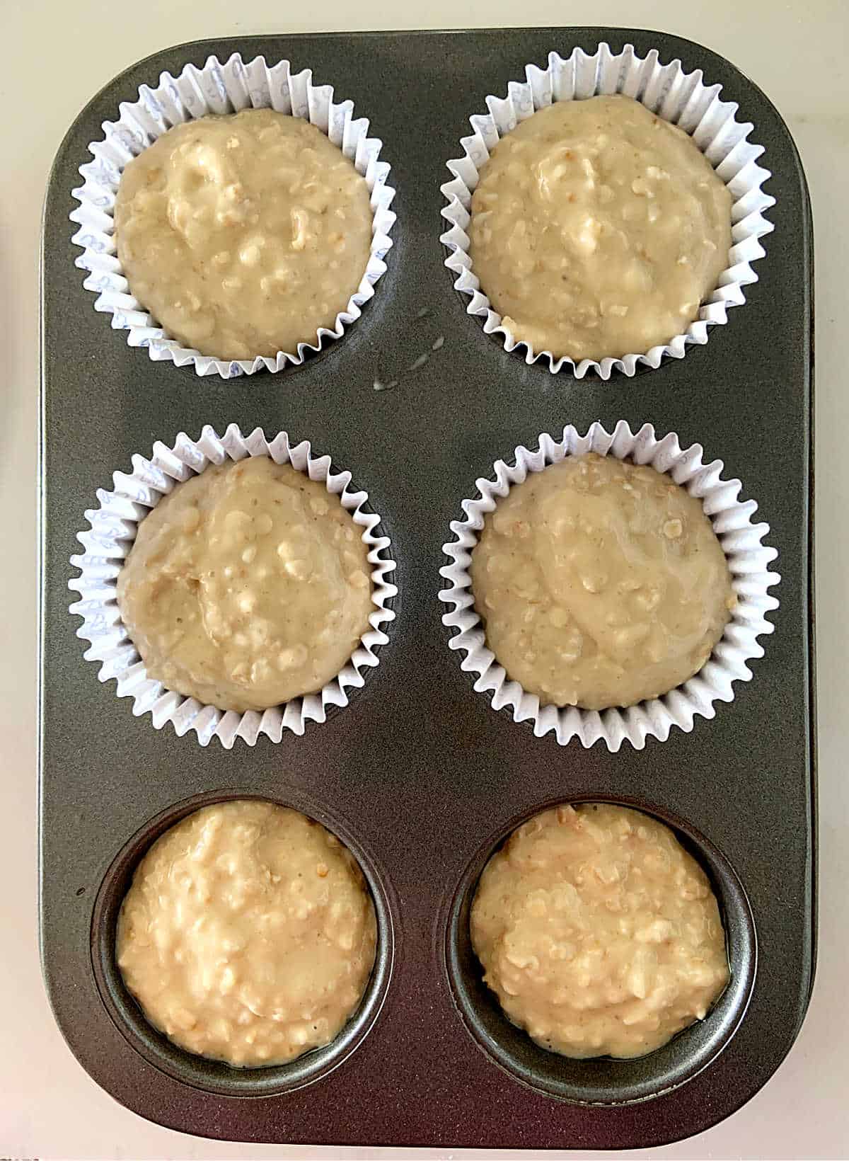 Dark 6-muffin metal pan filled with muffin batter; four have paper liners and two don't