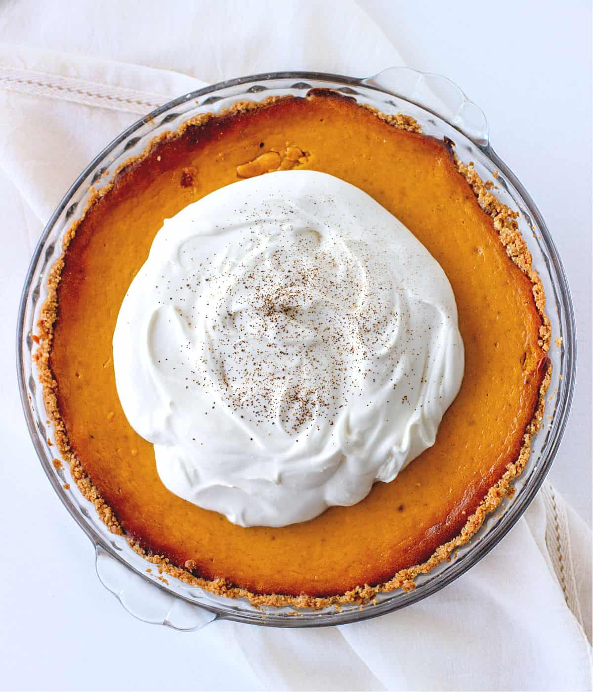 Top view of whole pumpkin pie with cream on top on a glass dish. White cloth and surface