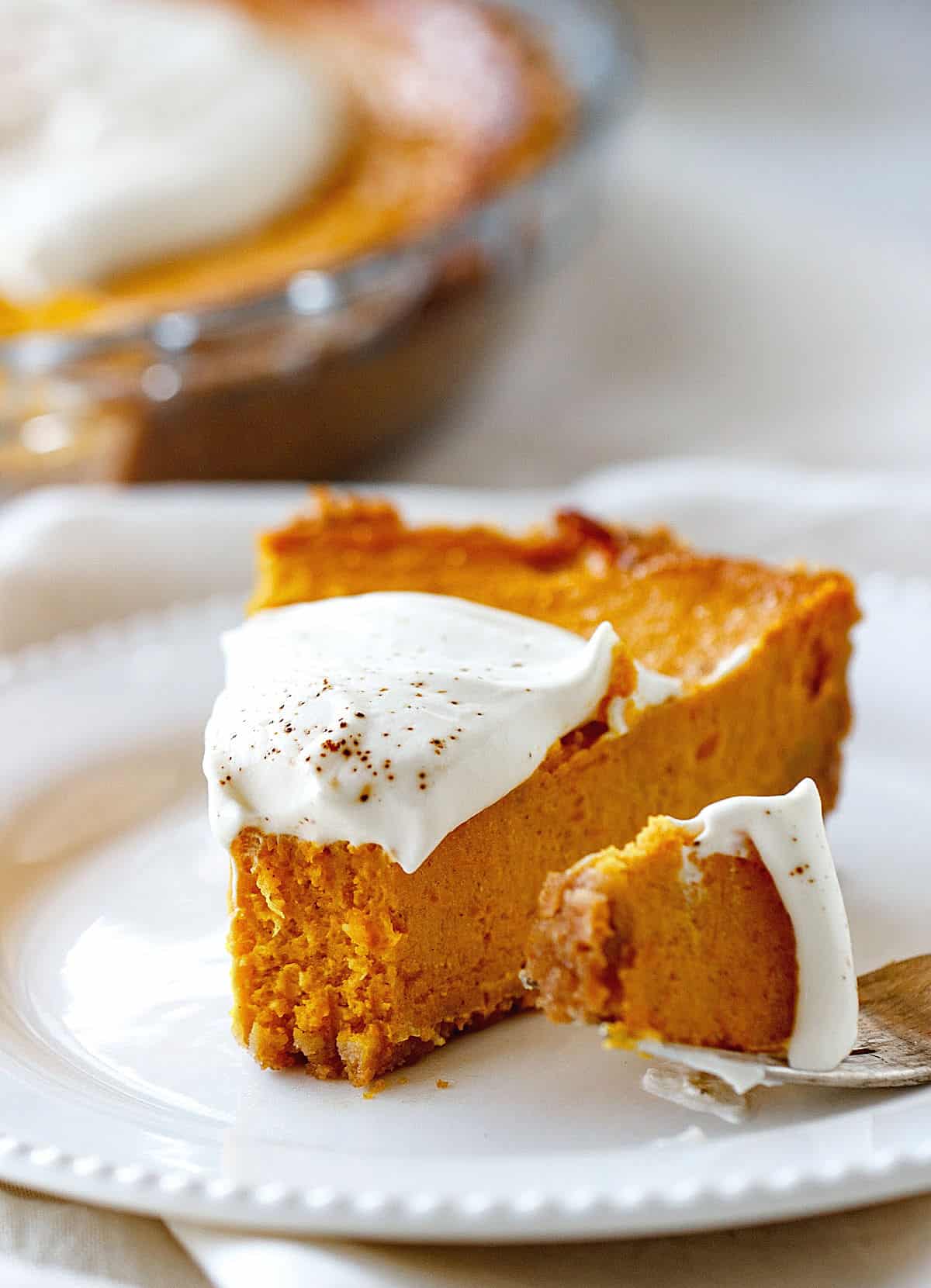A single slice of pumpkin pie with cream on top on a white plate, a fork with a bit beside it