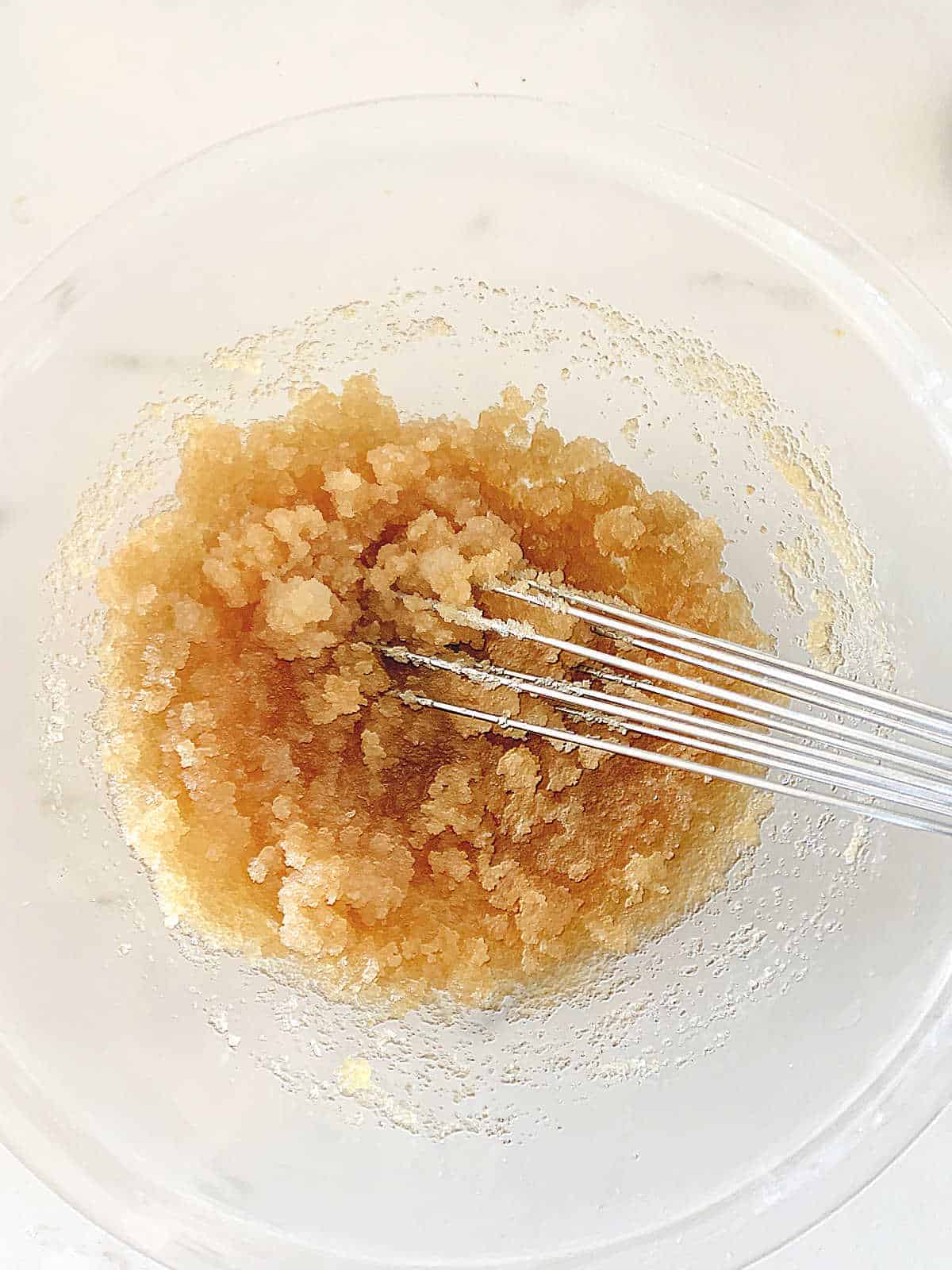 Glass bowl with oil and sugar being mixed with a whisk.