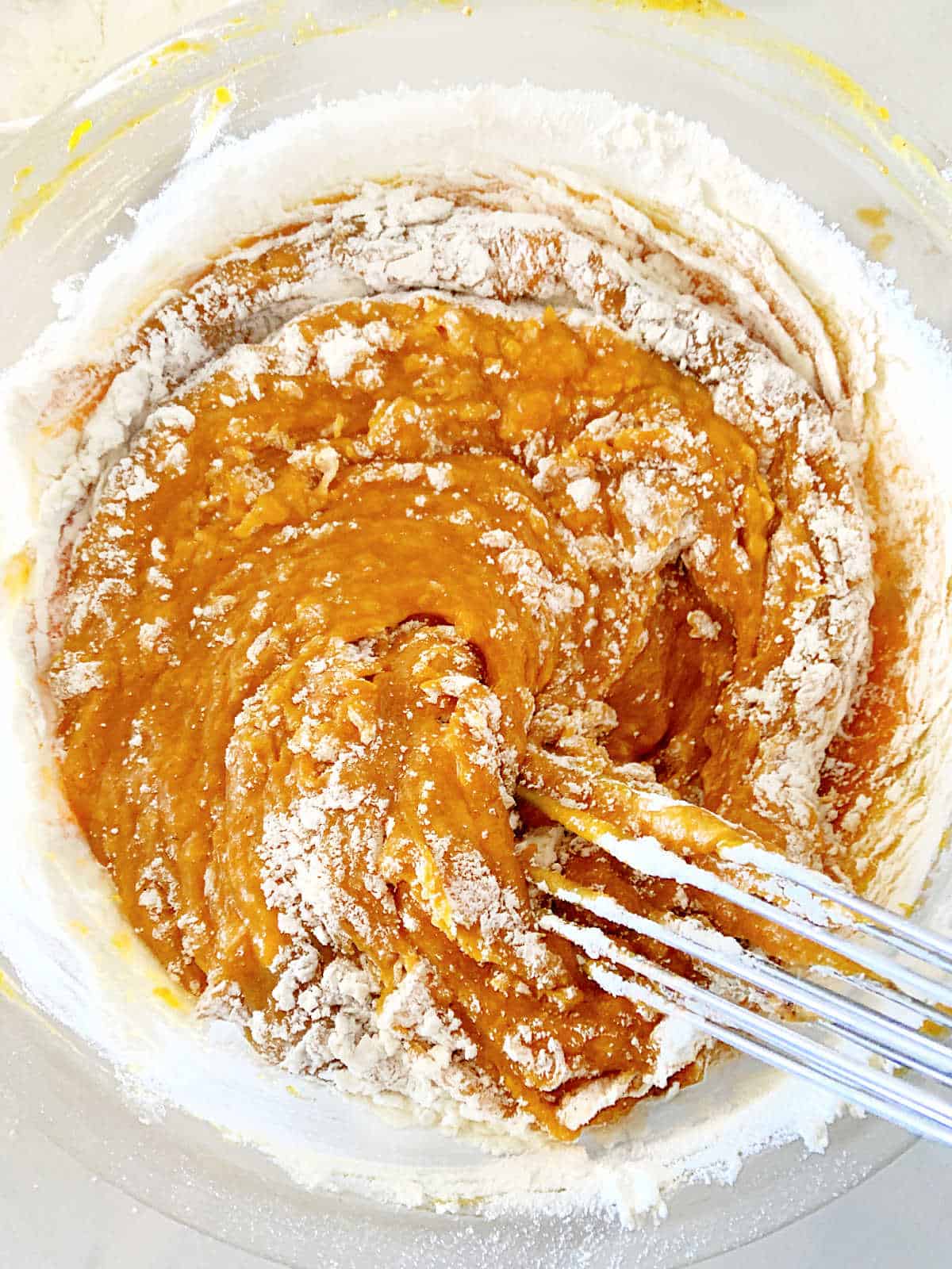 Close up of flour added to pumpkin batter in a glass bowl. A whisk.