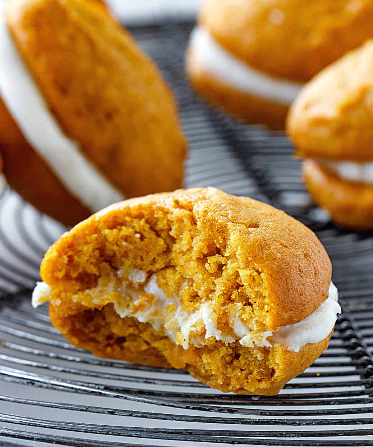 Close-up of bitten pumpkin whoopie pie on a dark wire rack, other whoopies blurred in background.