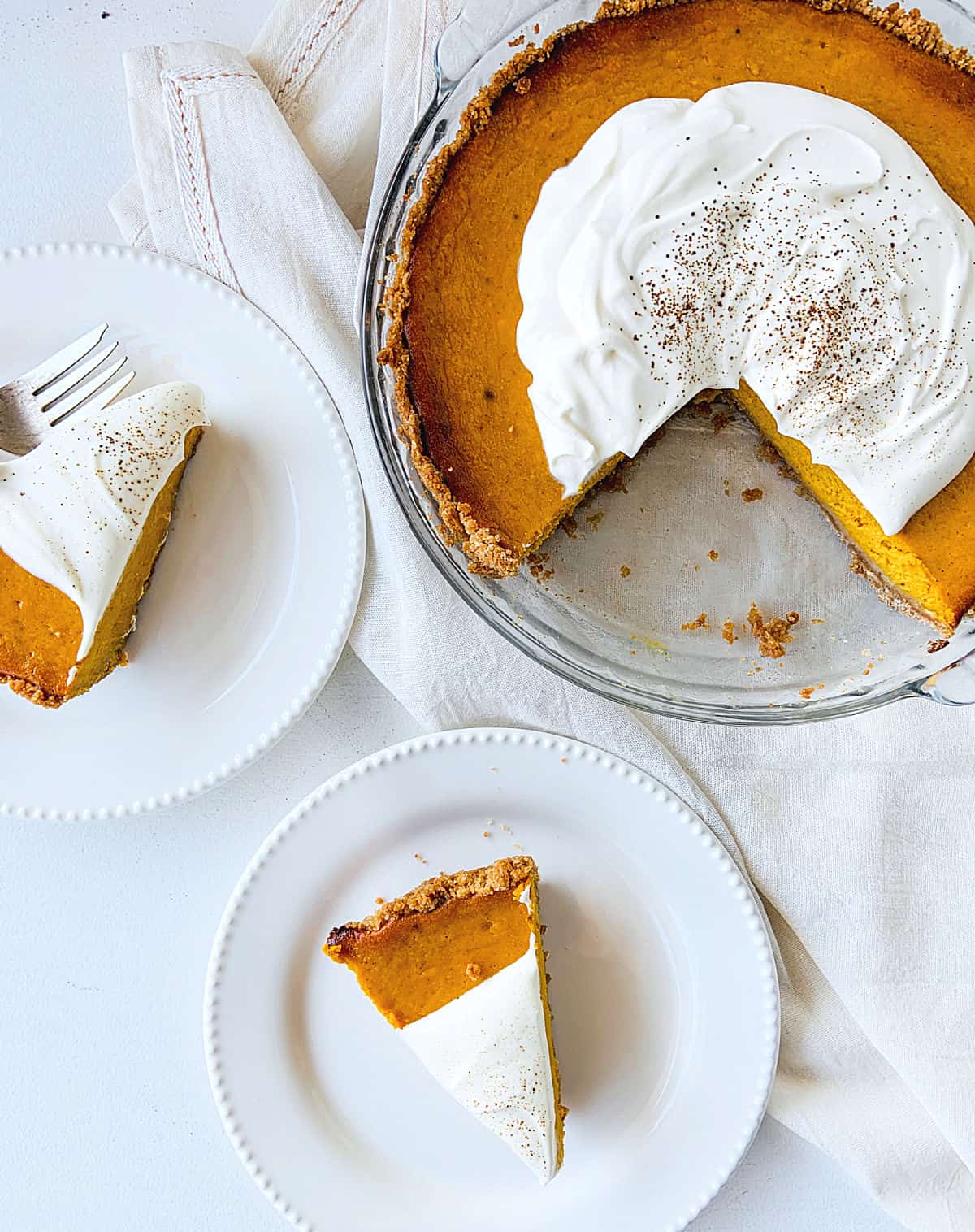 White plates with pumpkin pie slices, rest of pie in glass dish on white cloth and table